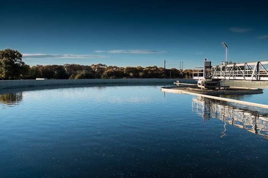 A large, circular water treatment tank reflecting the clear blue sky, nestled amid greenery and industrial structures at the Southside Water Reclamation Plant in Albuquerque. The setting suggests a calm day with no visible human activity.