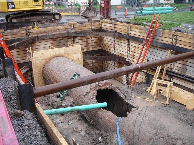 The construction site at the Buffalo Sewer Authority reveals a large, broken brick sewage pipe in an excavation pit. Supported by wooden panels and equipped with ladders for access, the area is scattered with various construction tools and materials, indicating ongoing efforts to enhance local sewage treatment.