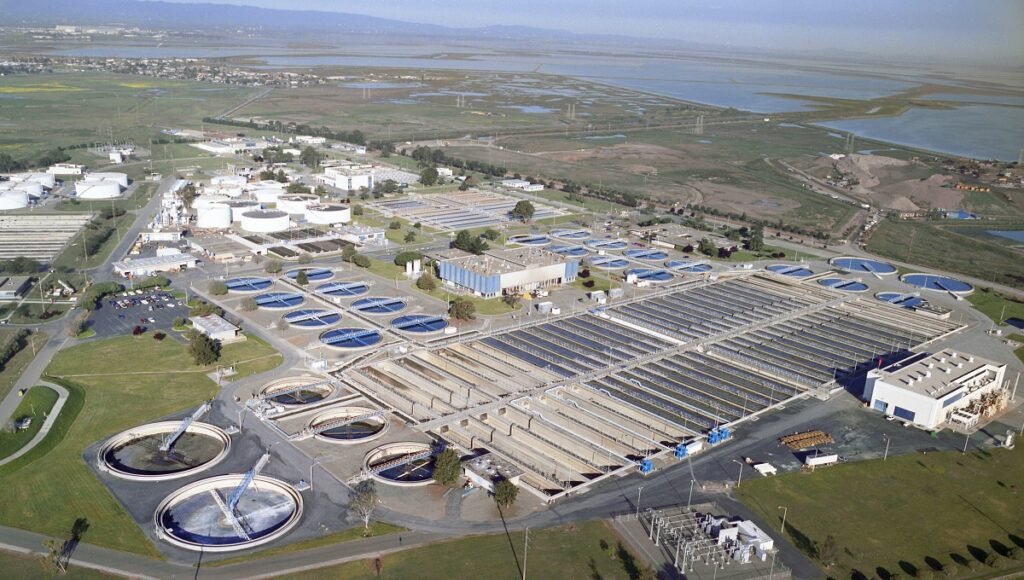 Aerial view of a sprawling wastewater treatment facility in San Jose, showcasing circular and rectangular tanks amidst lush greenery and nearby marshlands. The site is dotted with buildings and storage tanks throughout the expansive complex.