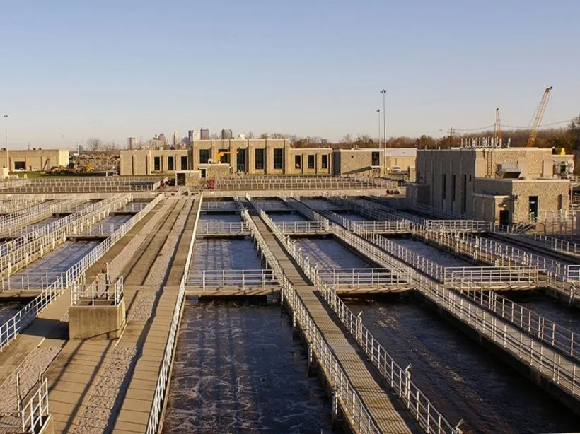 Aerial view of the Columbus Jackson Pike wastewater treatment plant reveals multiple rectangular concrete basins, walkways, and railings. Buildings and cranes stand in the background under a clear sky, showcasing an impressive infrastructure that keeps the sewers running efficiently.