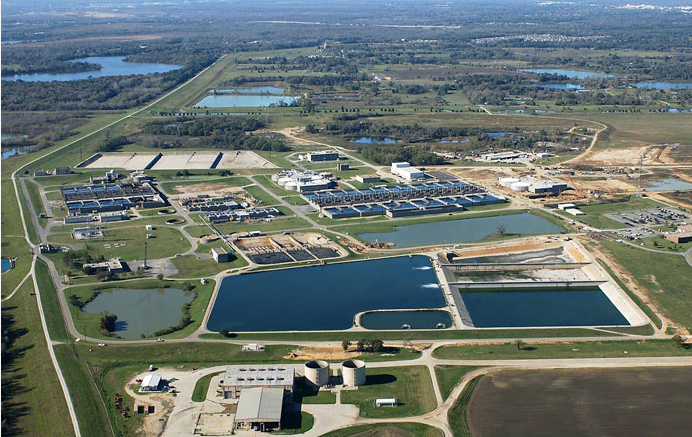 Aerial view of a wastewater treatment plant on the Southside, featuring large rectangular and circular water tanks. Open fields and trees surround the facility, with a distant Dallas cityscape and water bodies in the background.