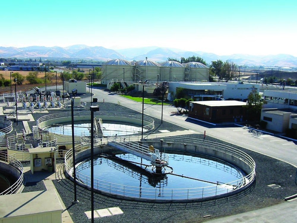 An aerial view of the Delta Diablo wastewater treatment plant reveals several circular tanks and surrounding buildings. The facility, managed by the Sanitation District, is set against hills under a clear blue sky, with pathways and utility structures visible around the tanks.