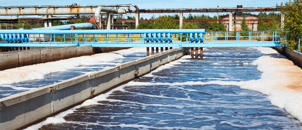 A wastewater treatment facility with concrete channels carries foamy, aerated water. Blue pipes run parallel to the channels. Industrial structures and greenery are visible in the background under a blue sky.