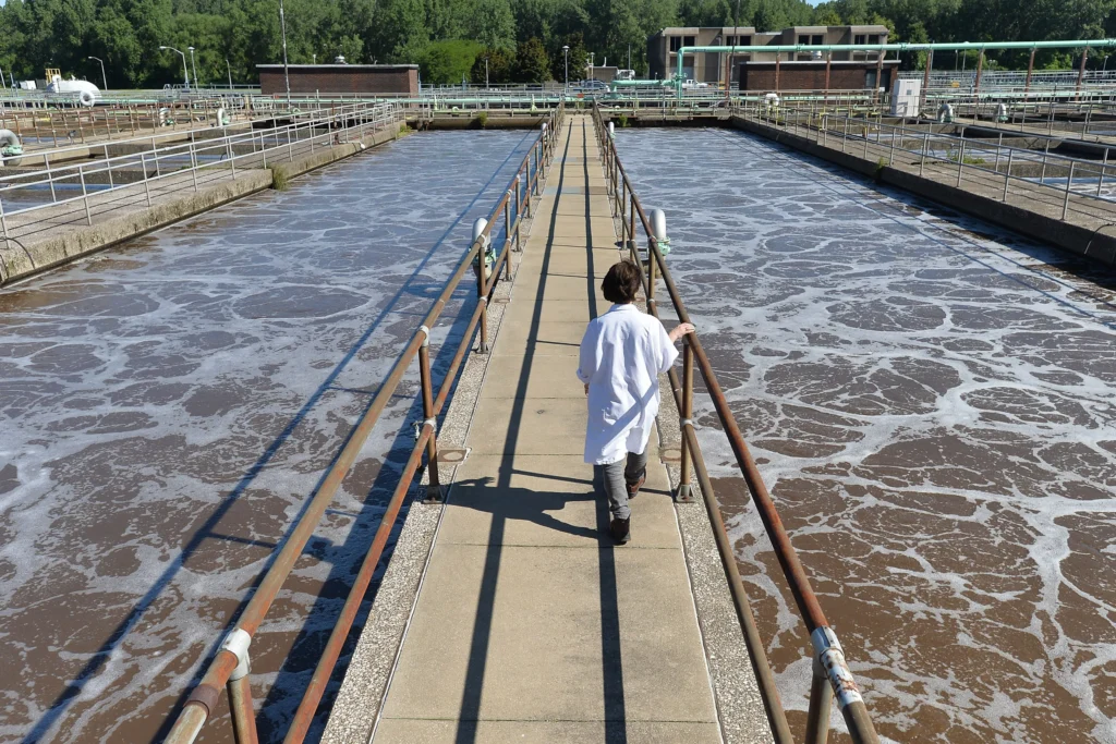 A person in a white lab coat walks on a narrow walkway over the sprawling Erie wastewater treatment plant, with aeration tanks on each side. The foamy, swirling water creates a contrast against the backdrop of trees and buildings.