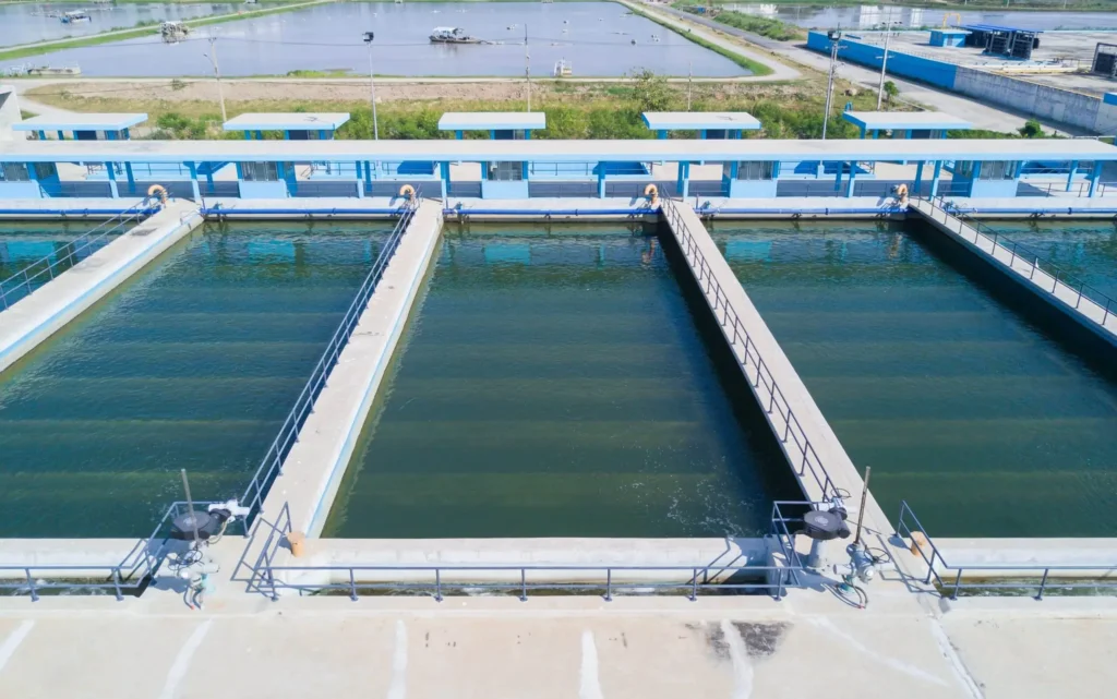 Aerial view of a rectangular wastewater treatment facility with three long, parallel filtration tanks separated by walkways. The background shows more water tanks and grassy areas under a clear sky.