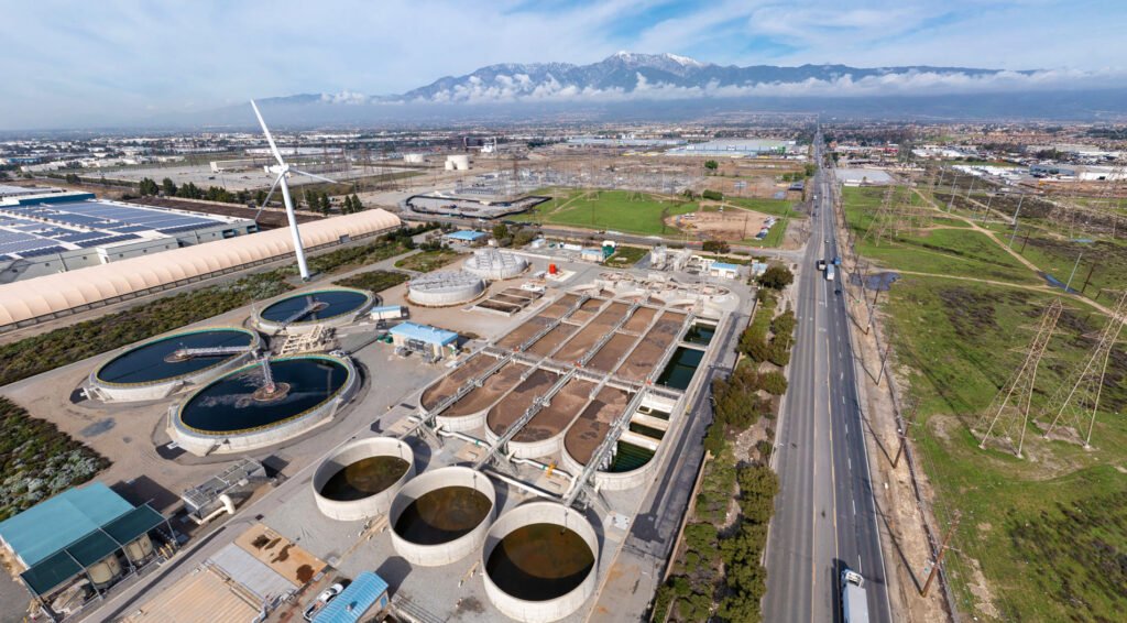 Aerial view of a regional water treatment facility with circular and rectangular tanks. A wind turbine from the Utilities Agency stands nearby. A road and power lines run alongside. In the distance, an Inland Empire cityscape and mountains are visible under a partly cloudy sky.