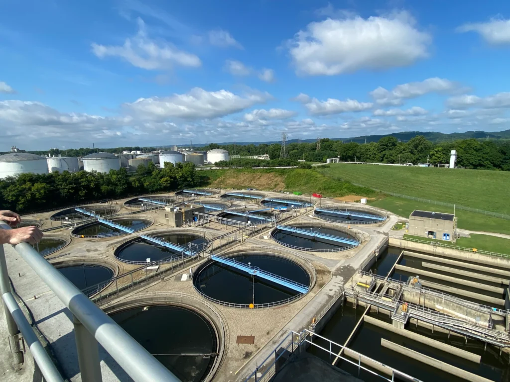 View of the Jefferson County Sewage Treatment Plant in Louisville, featuring multiple circular tanks surrounded by lush greenery. Silos and infrastructure stand in the background under a partly cloudy blue sky, with a railing prominently in the foreground.