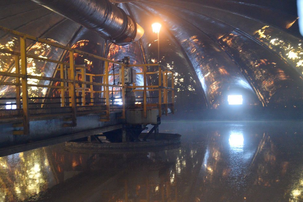 Interior of an industrial dome with reflective surfaces, yellow railings, and overhead pipes, illuminated by artificial light.