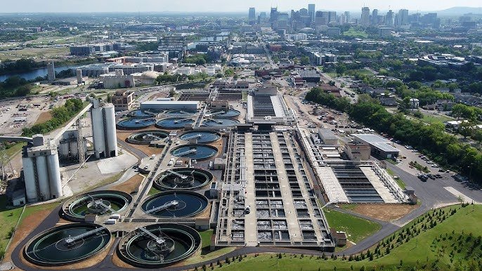 Aerial view of the Nashville Central Wastewater Treatment Plant with multiple circular basins and rectangular structures. The facility is situated near a river, with the city skyline visible in the background under a clear sky.