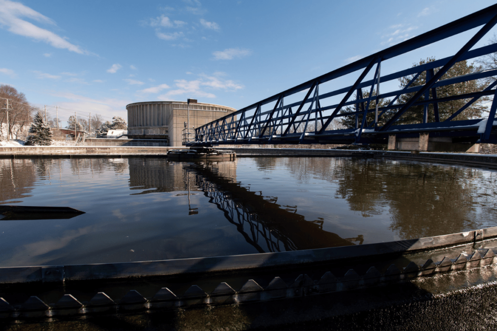 A large, circular water treatment tank reflects the blue sky, embodying the essence of water pollution control. A metal bridge spans over the tank, while an industrial building and trees are visible in the background. Shadows of railings add depth to this vital sewage treatment plant scene.