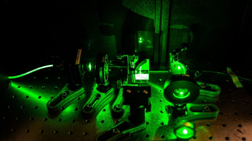 A complex assembly of optical equipment with lenses and mirrors is bathed in green laser light, enhanced by quantum precision. The setup rests on a perforated metal surface, creating a vivid and futuristic appearance.