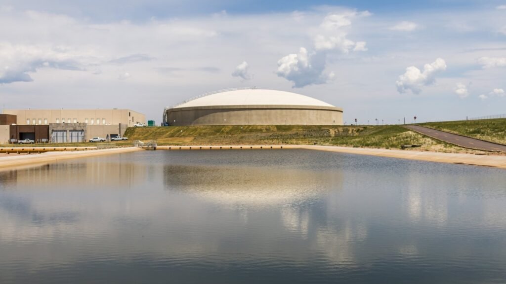 A large, round building with a white dome roof stands near a calm body of water, reflecting the structure. In the background, grassy fields stretch beneath a cloudy sky. Nearby, Colorado Springs Utilities' wastewater treatment plant and parked cars complete the serene landscape.