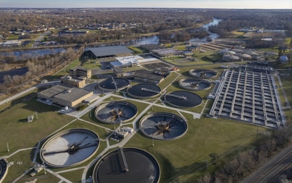 Aerial view of a large wastewater treatment facility, featuring multiple circular and rectangular tanks. The plant, designed with cost-effective solutions, is surrounded by grassy areas, roads, and a river in the background. Residential neighborhoods are visible in the distance.