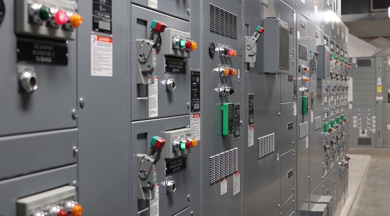 A corridor of industrial control panels with various switches, buttons, and dials. The metallic gray panels feature red, green, and orange indicators. Labels hint at energy consumption metrics in a technical setting reminiscent of a wastewater treatment plant.