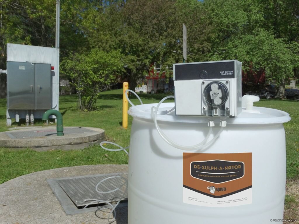 A white barrel labeled "DE-SULPH-A-NATOR" is equipped with a fluid pump machine on top. It stands on a concrete platform in a grassy area, efficiently handling hydrogen sulfide in wastewater, with trees and utility boxes in the background.