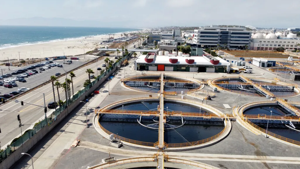 Aerial view of the expansive Hyperion Wastewater Treatment Plant near a beach. Several circular tanks are visible, surrounded by buildings and a parking lot. The coastline with waves and sandy beach extends into the distance on the left.