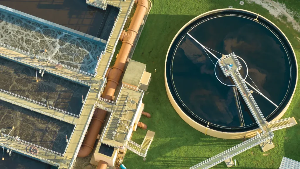 Aerial view of a wastewater treatment plant reveals rectangular aeration tanks on the left, swirling water in view, and a large circular sedimentation tank on the right, all surrounded by lush green grass. Structure details and pathways are clearly visible for those who live near.