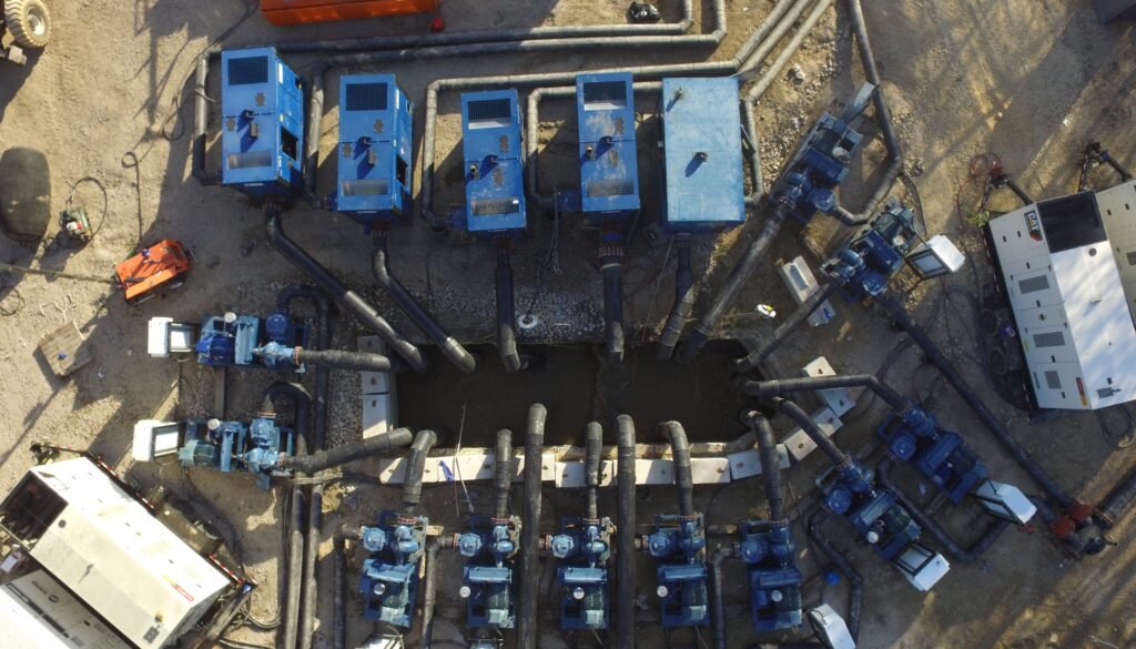 Aerial view of a wastewater treatment plant featuring multiple blue machinery units arranged around a rectangular pit. Large black hoses and various cables connect the machines, set amidst dirt and additional equipment. This industrial setup near Leon Creek operates with precision and care.