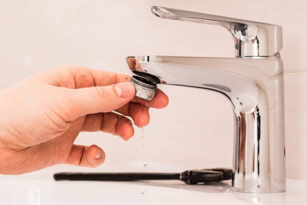 A hand carefully unscrews the aerator from a shiny chrome faucet, water droplets glistening in the light. A black wrench rests on the white surface, ready to help remove sediment and ensure your water flows smoothly.