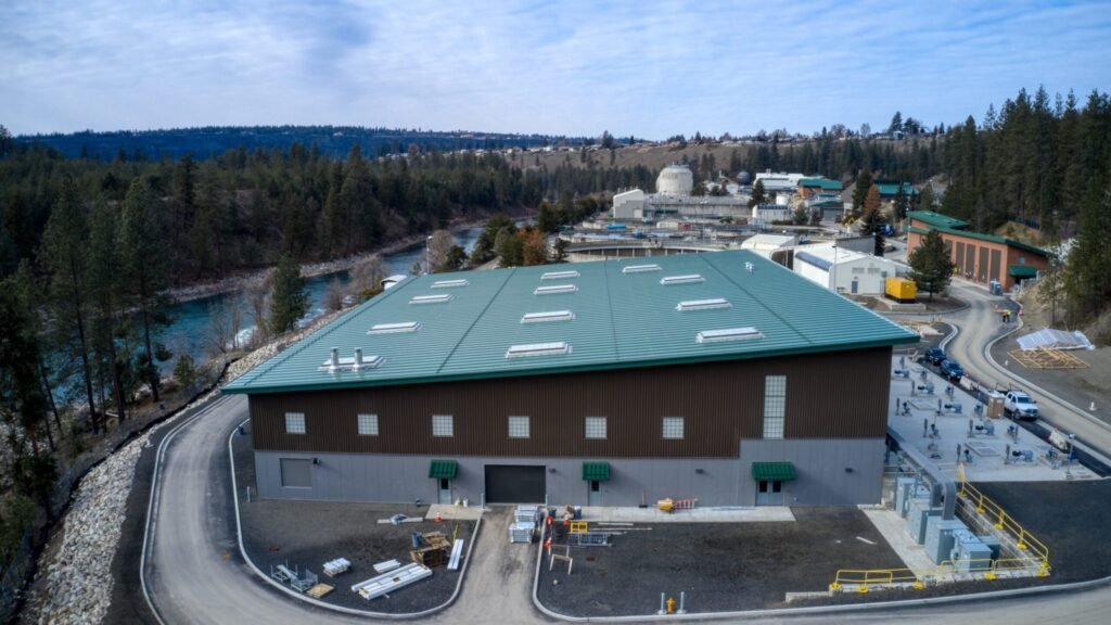 Aerial view of an industrial facility in Spokane, featuring a large green-roofed building. The site, near Riverside Park, is surrounded by trees and a nearby river. Overcast sky looms overhead while various equipment and materials dot the open areas around this water reclamation facility.