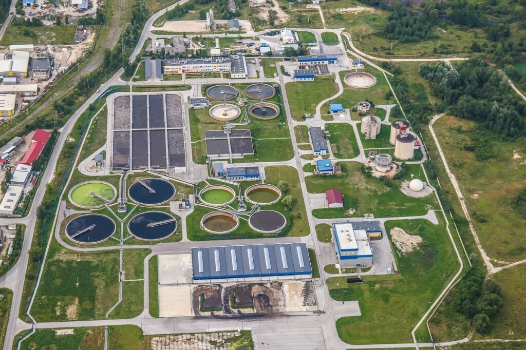 Aerial view of a wastewater treatment plant with multiple circular tanks, buildings, paved roads, and surrounding greenery.