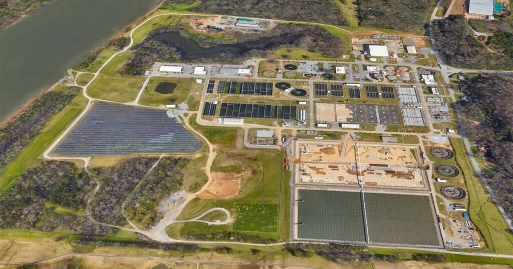 Aerial view of a large wastewater treatment facility at Moccasin Bend, nestled beside a river. The complex features numerous rectangular basins, buildings, and open areas. Surrounded by green fields and patches of trees, the treatment plant seamlessly blends with its natural environment.