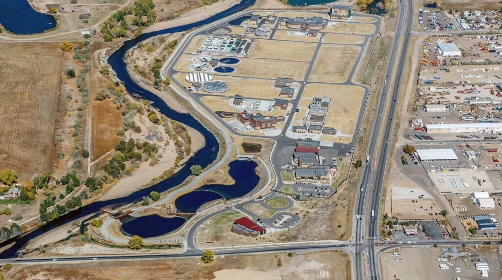 Aerial view of a wastewater treatment plant bordered by a river on one side and a road on the other. The facility includes several buildings, tanks, and rectangular sections. Surrounding areas feature industrial buildings and scattered trees.