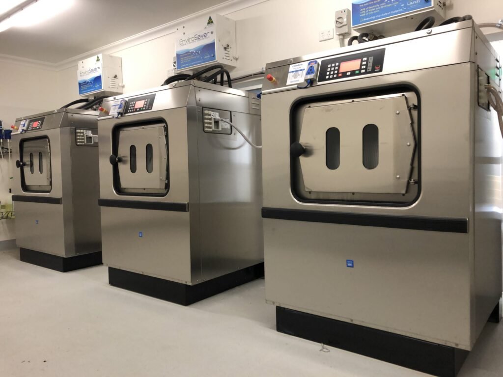 Three industrial washing machines equipped with an ozone laundry system are lined up in a clean, well-lit laundry room. Each machine features a digital control panel and a front-loading door with a rectangular window. The light-colored floor and support equipment on the walls complete this efficient setup.