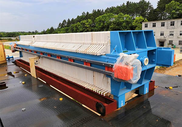 A large industrial filter press with a blue and white frame is situated outdoors on a black platform. Trees and a building are visible in the background. The machine, showcasing its plate and frame design, is wrapped in some protective plastic material, illustrating its robust working principle.