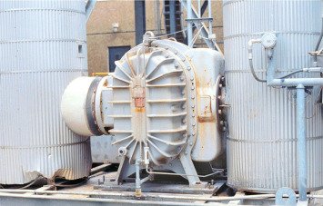 An industrial air compressor with a large, circular fan wheel sits between two tall, cylindrical tanks. The metal components show signs of wear and rust, resembling those on an aging centrifugal blower. In the background, a building with a small window adds an industrial vibe.