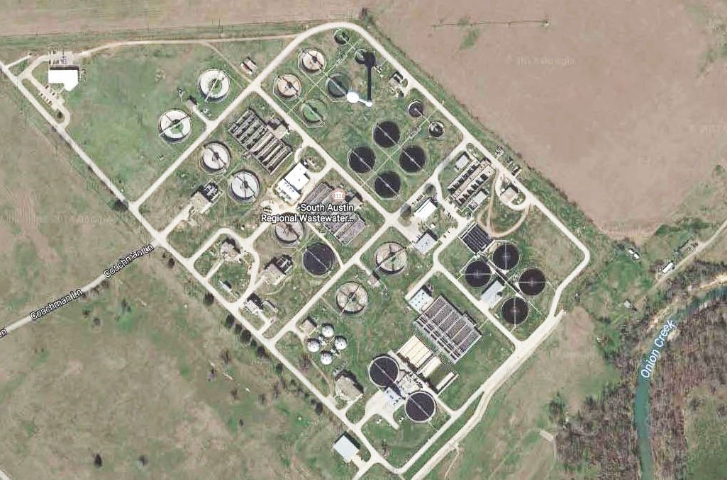 Aerial view of the South Austin Regional Wastewater Treatment Plant. The facility features numerous circular and rectangular tanks, interconnected buildings, and service roads. Onion Creek is visible on the right side, bordered by grass and fields.