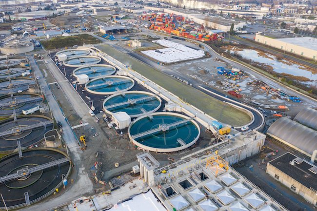 Aerial view of a large wastewater treatment plant featuring several circular clarifiers. The intricate stages of wastewater treatment unfold amidst an industrial area with buildings, construction equipment, and shipping containers nearby. Paved roads and green spaces provide a structured contrast.