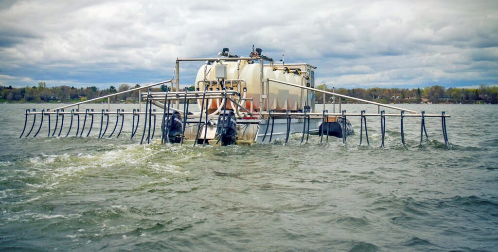 A large watercraft with multiple tanks and pipes maneuvers on the surface of a lake, stirring the water in its path. Designed for wastewater treatment, it efficiently disperses alum. The sky is overcast, with trees and houses visible along the distant shoreline.