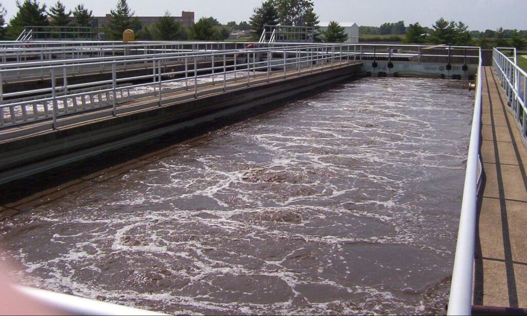 The aeration tank at the wastewater treatment plant showcases frothy water, indicating active treatment. Metal railings border the tank, while trees stand in the background under a clear sky. Nearby lies the anoxic zone, crucial for managing nutrient removal.