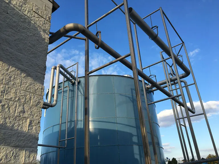 A large blue industrial tank, part of the wastewater treatment system, is surrounded by metal pipes and a sturdy framework. It stands next to a building with a rough-textured, light-colored wall. The clear blue sky above has a few clouds drifting by.