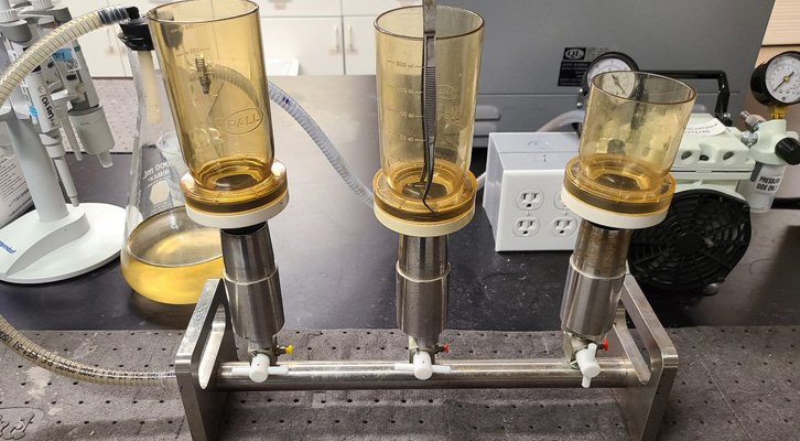 A laboratory filtration setup with three filtration funnels, each with a transparent yellow vessel on a metal stand, is used in wastewater treatment. Tubing is attached, and various lab equipment is visible in the background, enhancing the precision of Mlvss analysis.