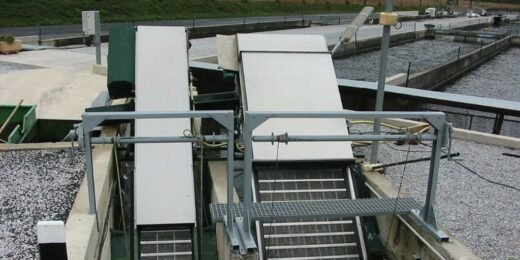 An industrial setting with two large, inclined conveyor belts near a body of water used for wastewater screening. The area is surrounded by metal railings and concrete surfaces, with additional equipment visible in the background.