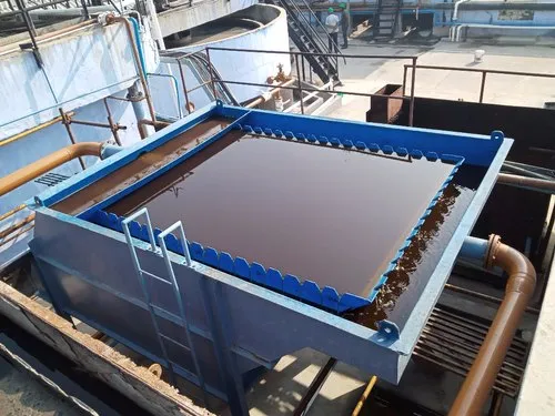 A view of an industrial oil-water separator featuring a large, rectangular blue tank and ladders on the side, surrounded by pipes. The separator is filled with a dark liquid, likely oil. In the background, you can see machinery alongside a lamella clarifier enhancing separation efficiency.