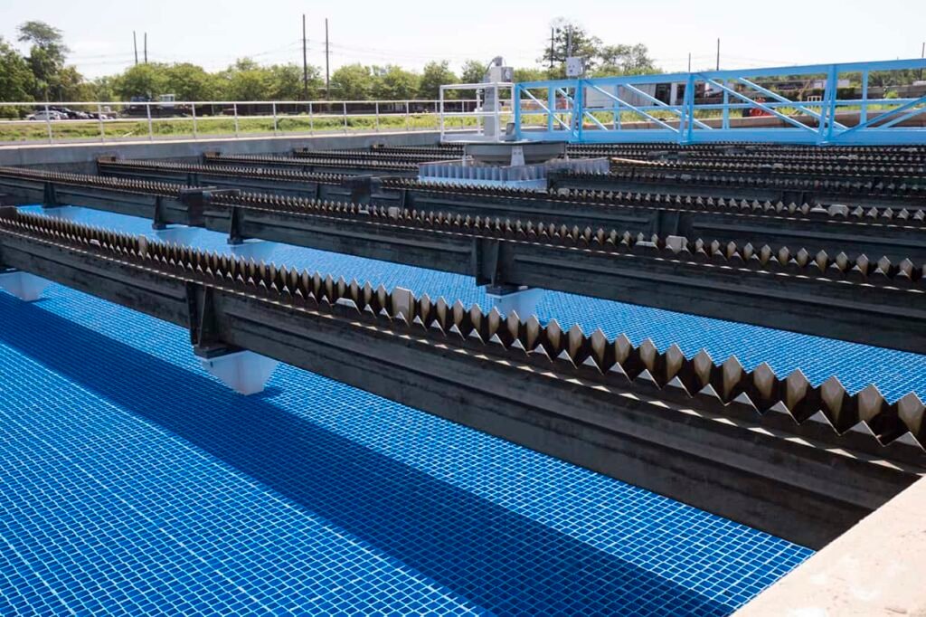 An image of a water treatment facility showcases several rectangular pools with blue-tiled bottoms, enhanced by advanced tube settlers. The pools are separated by evenly spaced metal beams with serrated edges. Fencing and trees provide a serene backdrop under the clear sky.