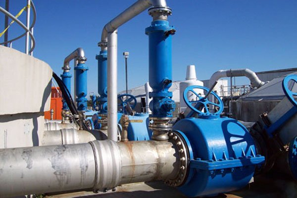 The industrial setting features a complex piping network with large metal pipes and blue valves, under a clear sky. This intricate system, part of the water treatment plant, includes numerous joints and flanges, set against structures typical of such facilities.