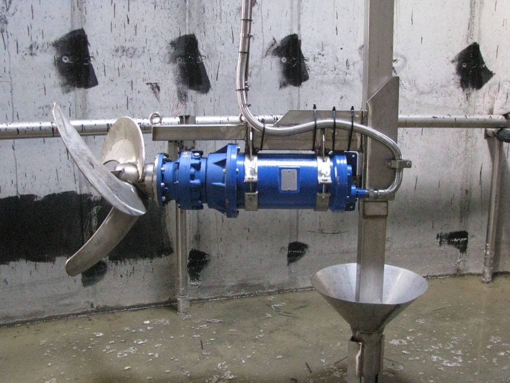 A blue industrial mixer, functioning as an agitator, with metal blades and connected pipes is mounted on a vertical metallic stand in a concrete-lined facility. The gray walls marked in black give a stark contrast to the wet floor, reflecting the efficient wastewater management process at work.