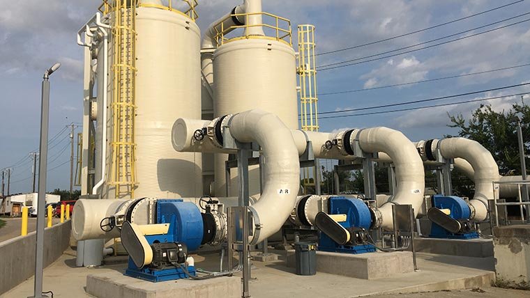 The wastewater treatment plant features large white cylindrical tanks and a network of interconnected piping. Blue machinery rests on concrete platforms in front of the tanks, while overhead power lines stretch across a partly cloudy sky, all working together to ensure efficient odor control.