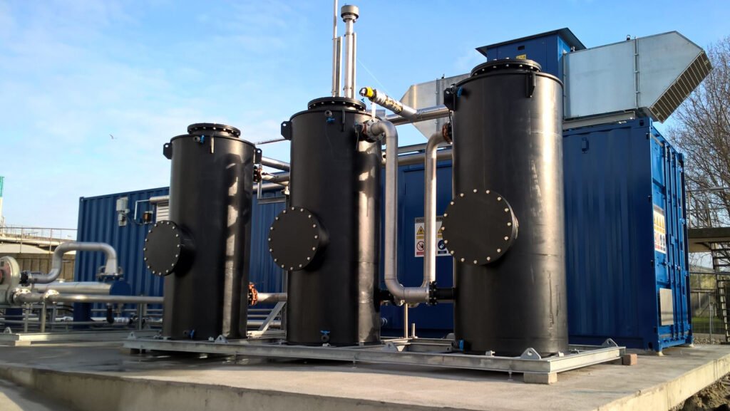 Three large black industrial tanks with pipes, next to a blue shipping container on a concrete platform.