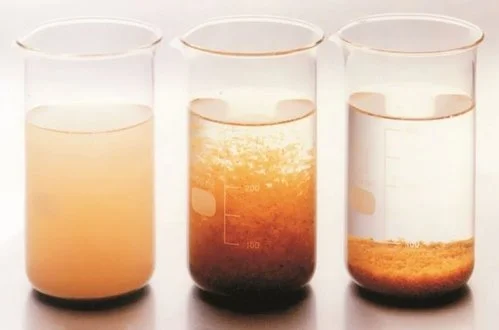 Three beakers are lined up, each containing wastewater with varying sediment levels. The first beaker is cloudy, the second has thick, suspended flocs, and the third shows sediment settled at the bottom with clearer liquid on top, demonstrating a stage of biological treatment.