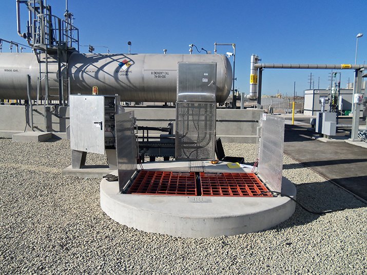 Industrial site with an open valve pit, red grate cover, and large cylindrical tanks in the background.