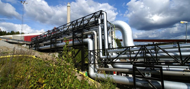 Industrial pipes under a cloudy sky beside a power station, surrounded by greenery and industrial buildings.