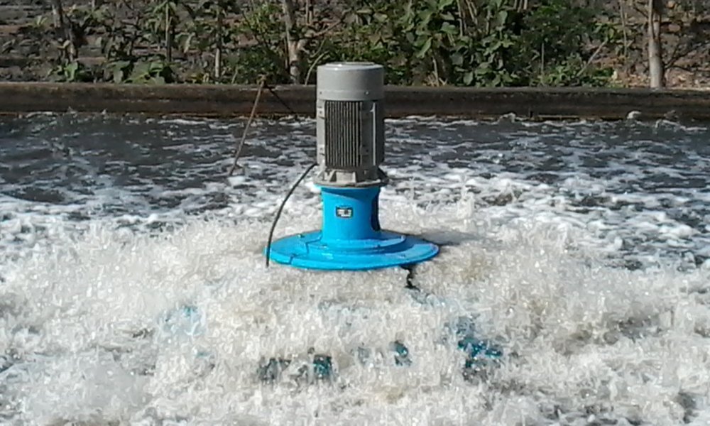 A blue submersible aerator churns water vigorously in the pool, its motorized action crucial for effective wastewater treatment. The surrounding area is bordered by concrete, with plants visible in the background as frothy waves ripple across the surface.