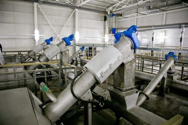 The image shows an industrial facility with large metallic machinery. Among the metal railings and structural beams under ceiling lights, several inclined conveyor systems for pollutant removal with blue motors are visible. The smooth concrete floor complements the efficient design.