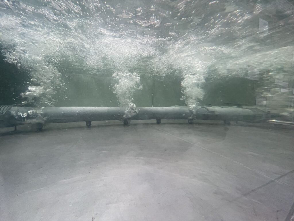 An underwater scene features a metallic pipe releasing streams of bubbles into clear water. These coarse bubble diffusers create a dynamic effect as bubbles rise and scatter against the backdrop of a concrete or grayish surface.