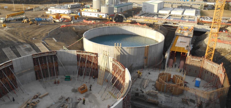 Aerial view of an innovative urban sanitation project featuring large circular structures. One is filled with water, while others are in various stages of construction. Surrounding buildings, materials, and equipment paint the scene of a cutting-edge concrete wastewater treatment plant in progress.